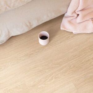 Quick-Step Alpha Bloom Pure Oak Blush vinyl flooring in a bedroom setting with soft pink textiles and a coffee cup