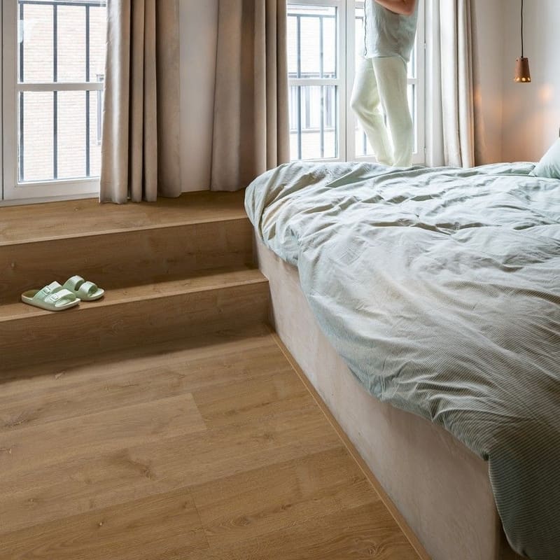 Bedroom installation of Quick-Step Alpha Bloom Elegant Oak Fumed vinyl flooring showing warm oak tones in a contemporary setting