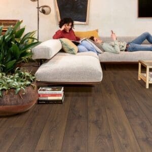 Quick-Step Peanut Brown Oak laminate flooring installed in a modern living room with people relaxing on a sofa