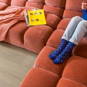 Person relaxing on coral sofa with Quick-Step Soft Oak Medium laminate flooring in contemporary living space